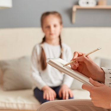 Psychologist taking notes while conducting session with female child, writing down important points about her emotional and mental condition, listening about little girl problems.
